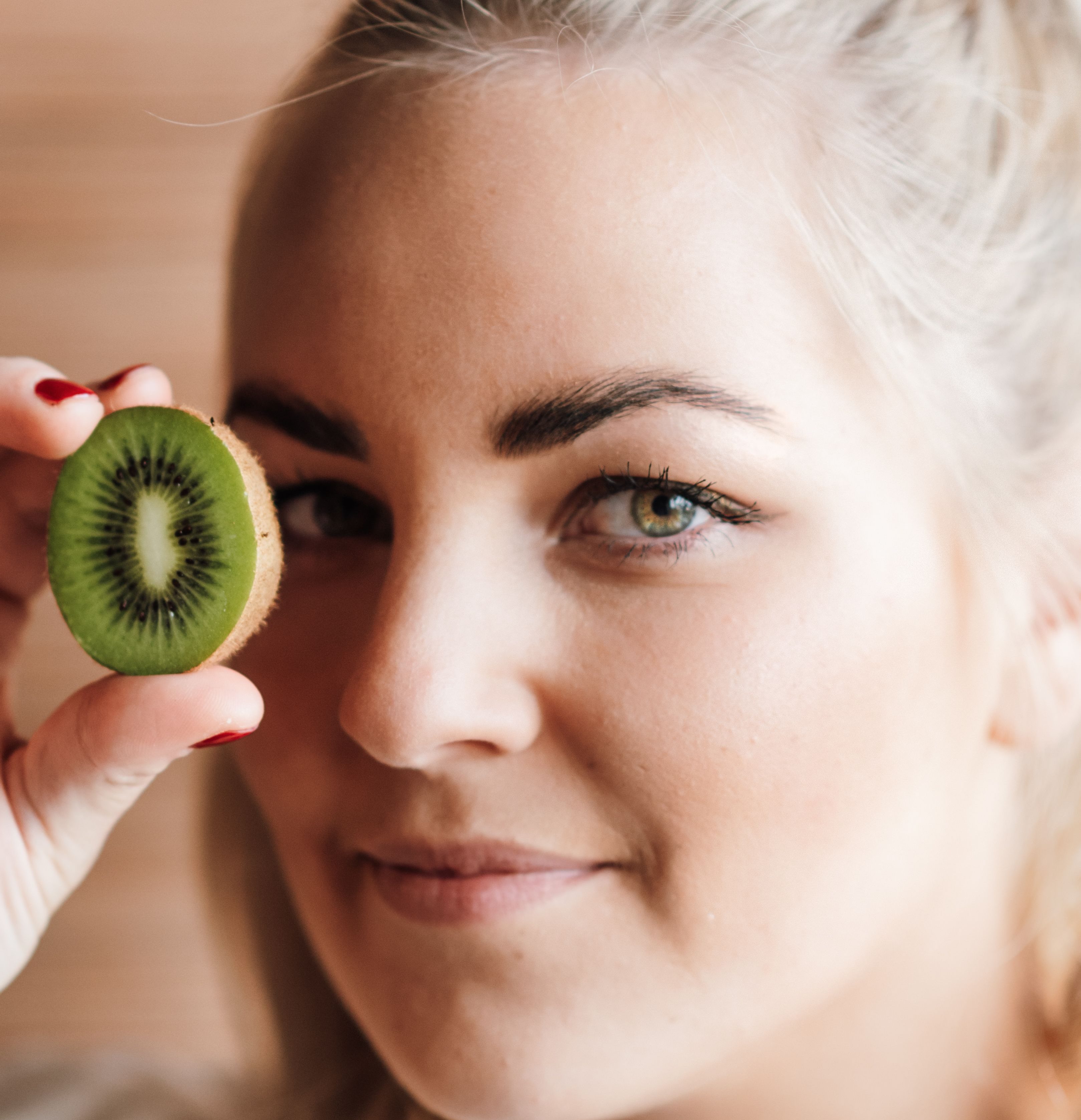 Young smiling female with half of ripe tasty kiwi near face looking at camera